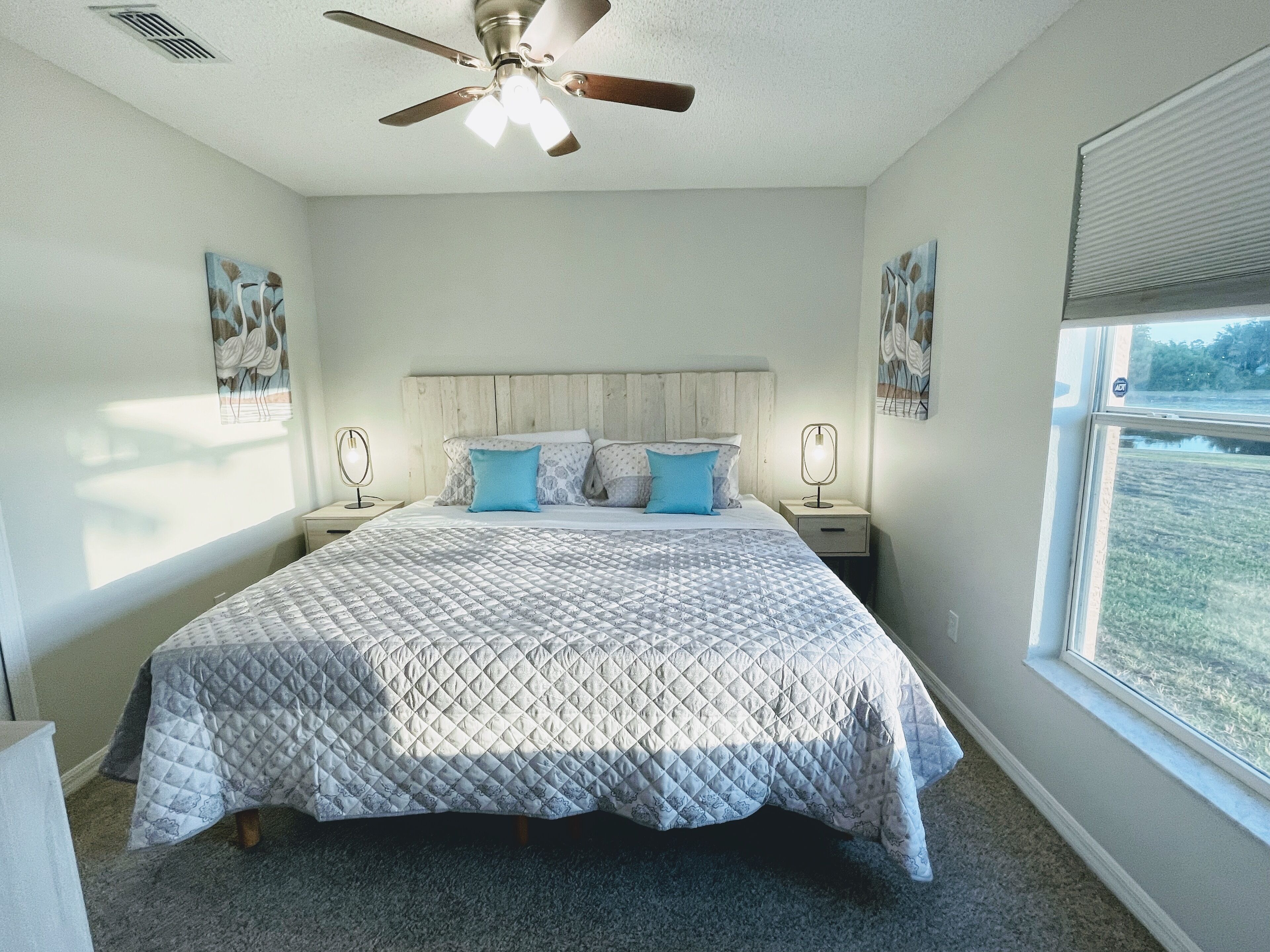 A bedroom with a bed, a ceiling fan, two bedside tables with lamps, and a window with a view of a grassy field.