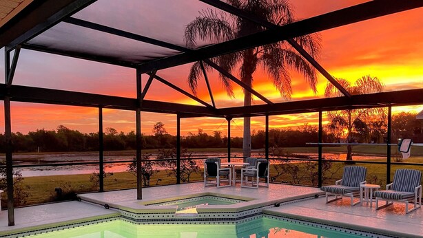 A covered outdoor pool area with a view of a sunset over a body of water, surrounded by chairs and a small table.