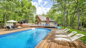 A swimming pool with wooden deck and lounge chairs, surrounded by trees and a house with a deck. - Contemporary retreat on 1.4 wooded acres with an open floorplan and private... (East Hampton)