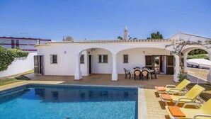 Uma área de piscina com piscina azul límpida, mesa e cadeiras, e um edifício branco com arcos.
