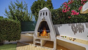 Uma área de churrasco ao ar livre com forno de pedra, um pátio com azulejos e vegetação ao redor.