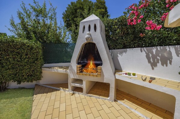 An outdoor barbecue area with a stone oven, a tiled patio, and surrounding greenery.