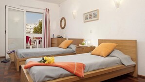 Quarto de hotel com duas camas individuais, cabeceira de madeira, uma mesinha, uma cadeira, janela com cortinas e luminária fixada na parede.