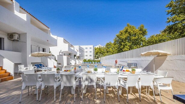 Uma área de refeições ao ar livre com mesas e cadeiras brancas, piscina ao fundo e um edifício branco com varandas.