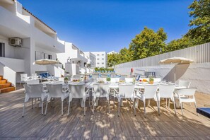 Un espace extérieur aménagé pour les repas, avec des tables et des chaises blanches, une piscine en arrière-plan et un bâtiment blanc doté de balcons.