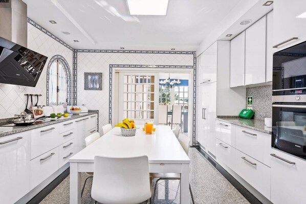 Een moderne keuken met witte kasten, een eettafel met stoelen, een groot raam en een Siemens koelkast.