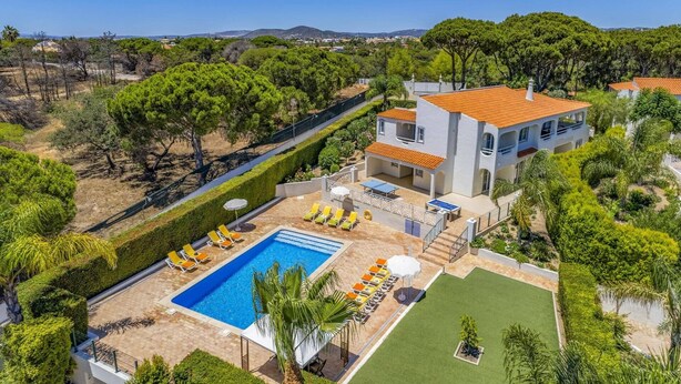 Una casa de dues plantes amb una piscina, envoltada de vegetació i amb turons al fons.