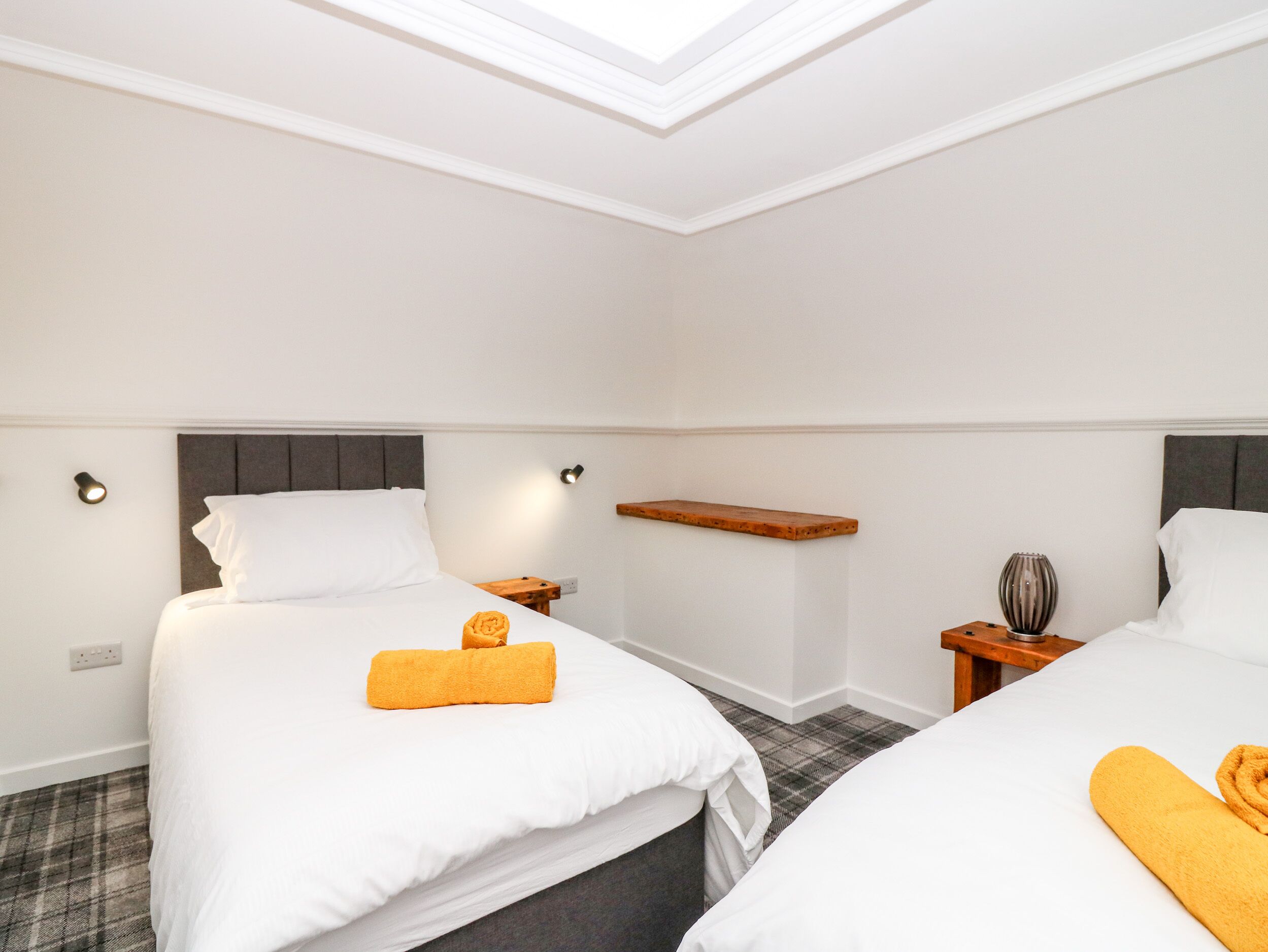 A hotel room with two beds, a skylight, a wooden shelf, and a small table with a vase.