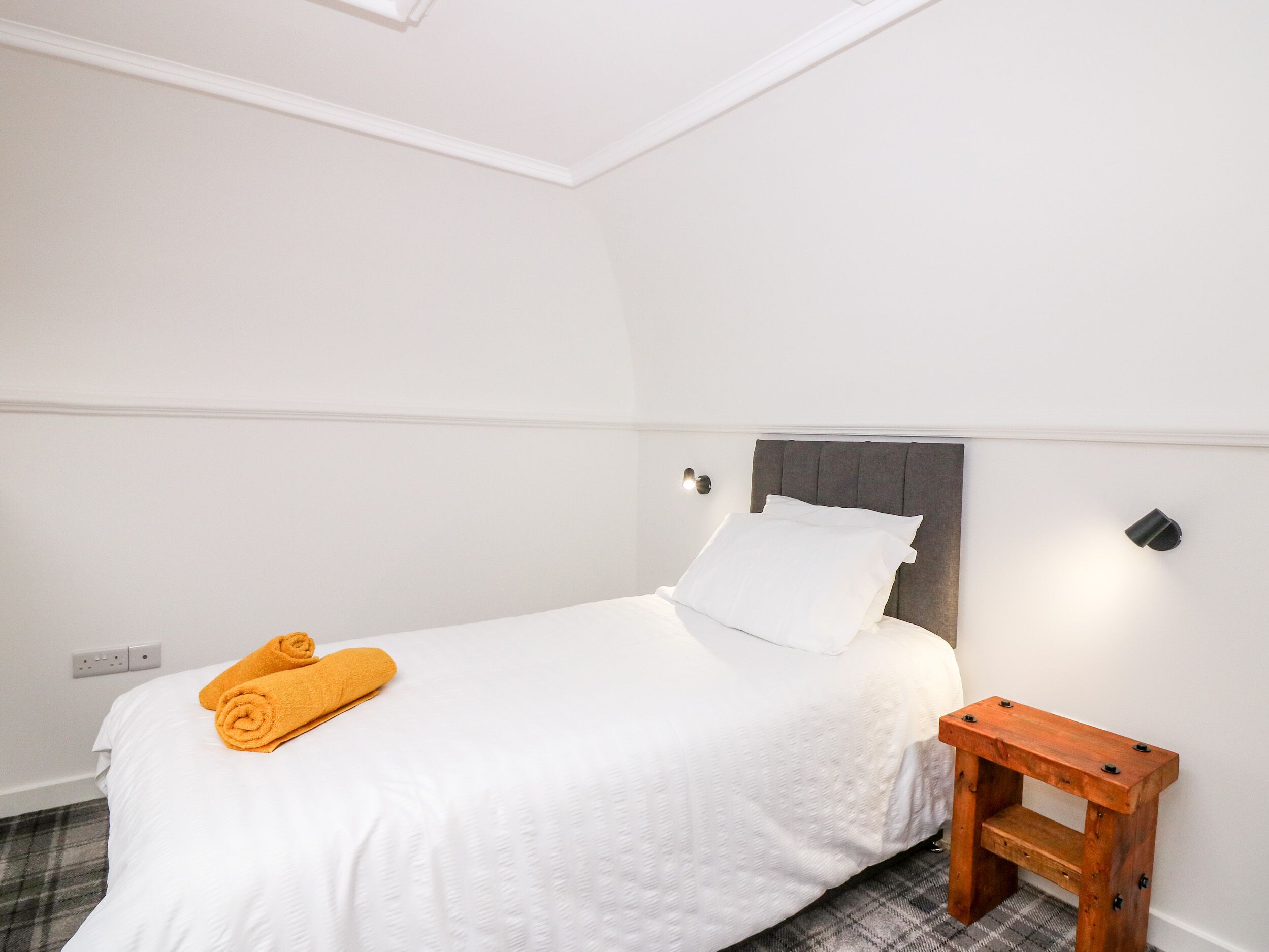 A neatly made bed with white bedding and a single white pillow, a small wooden nightstand beside it, and a pair of rolled towels on the bed.