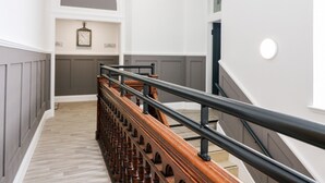 A staircase with a wooden handrail and a black metal handrail, leading to a landing with a clock and a small shelf. - The Court Room at The Institute Executive Apartments (Keith)