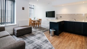 A modern kitchen with a dining area, a TV mounted above the counter, and a large window with blinds. - The Court Room at The Institute Executive Apartments (Keith)