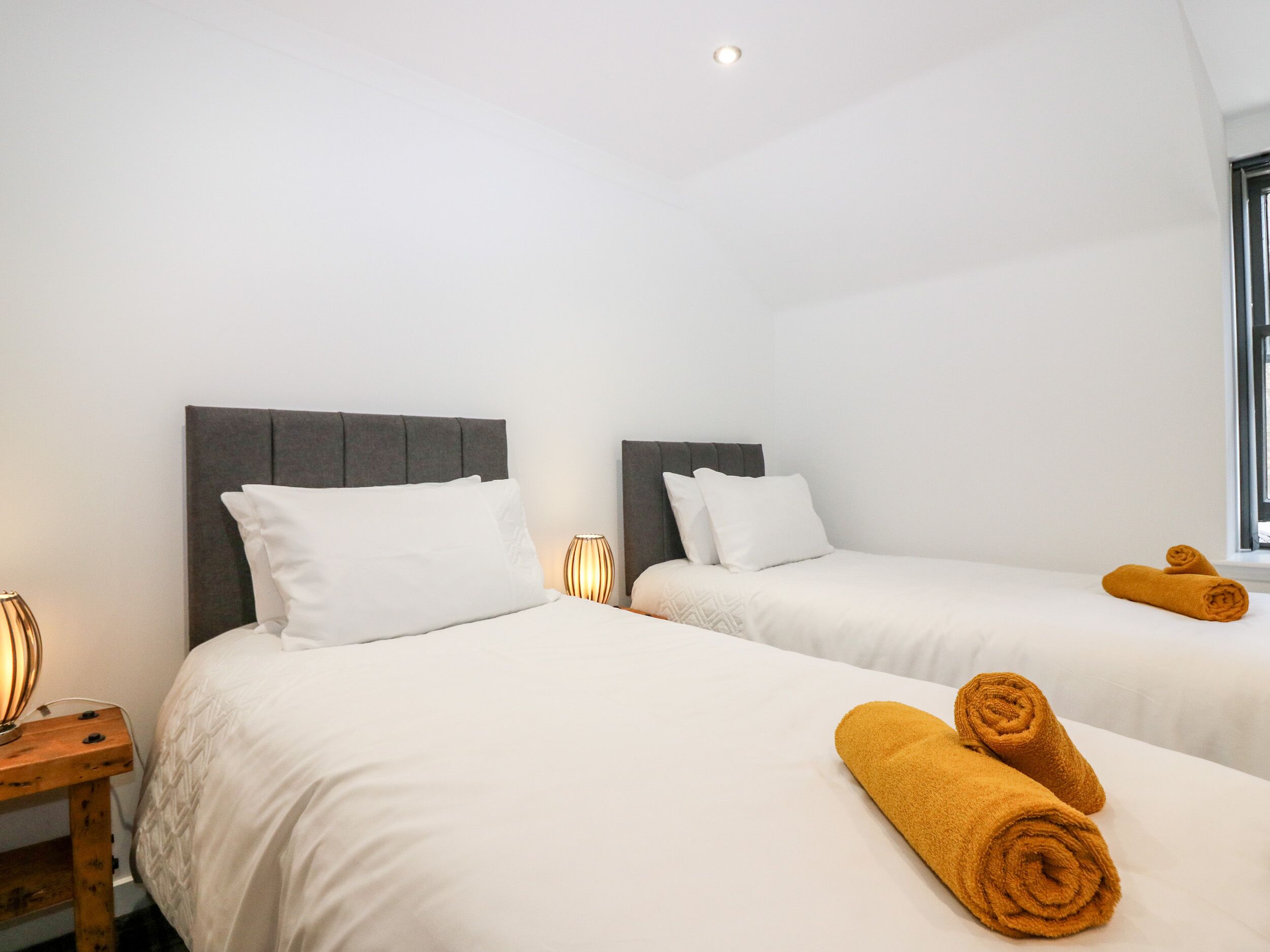 A hotel room with two beds, each with a grey headboard and white bedding. There are two bedside lamps and a window with a view.