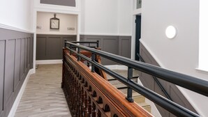 Eine Treppe mit hölzernem Geländer und einem schwarzen Metallgeländer, die zu einer Zwischenebene mit einer Uhr und einer wandmontierten Leuchte führt.