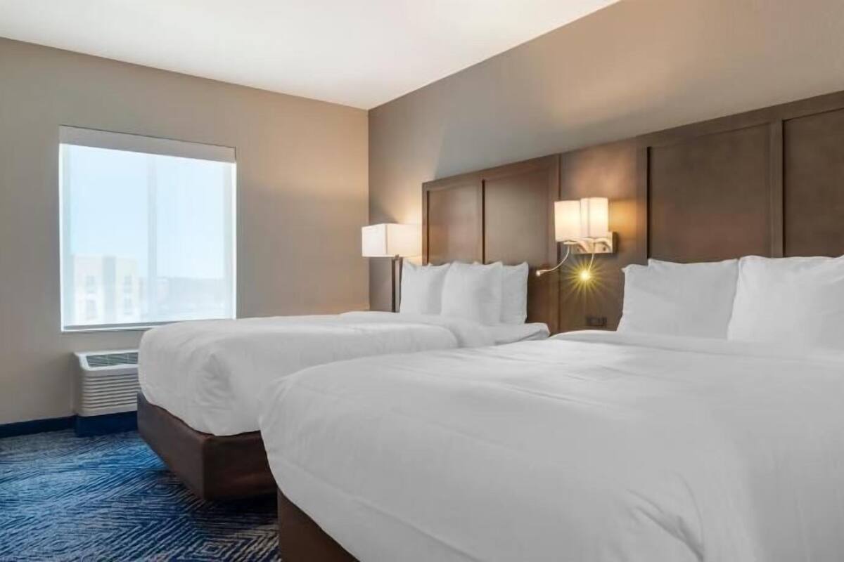 A hotel room with two beds, a large window, and a wall-mounted lamp.