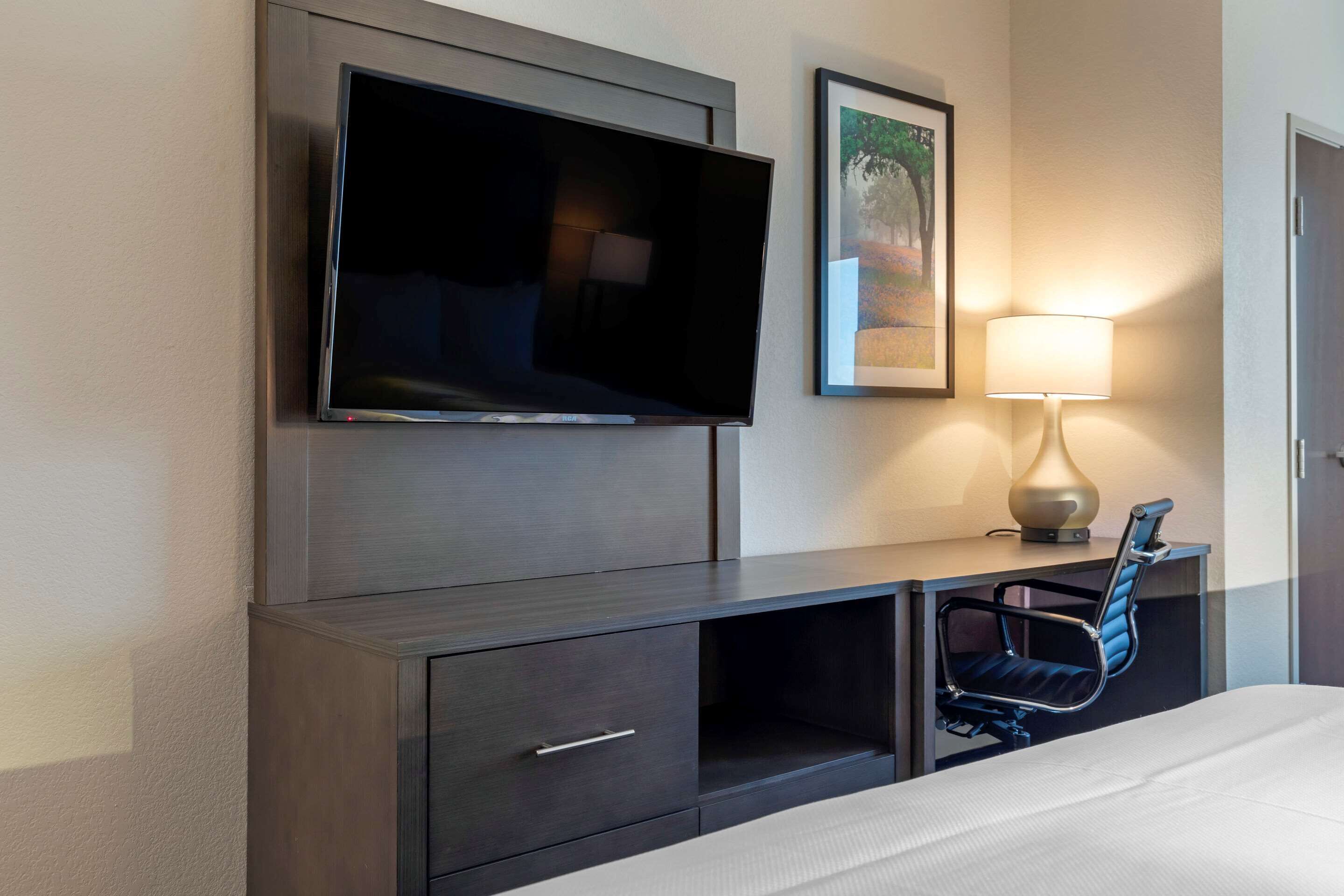 Habitación de hotel con televisión de pantalla plana montada en la pared, escritorio con silla, lámpara y cama.