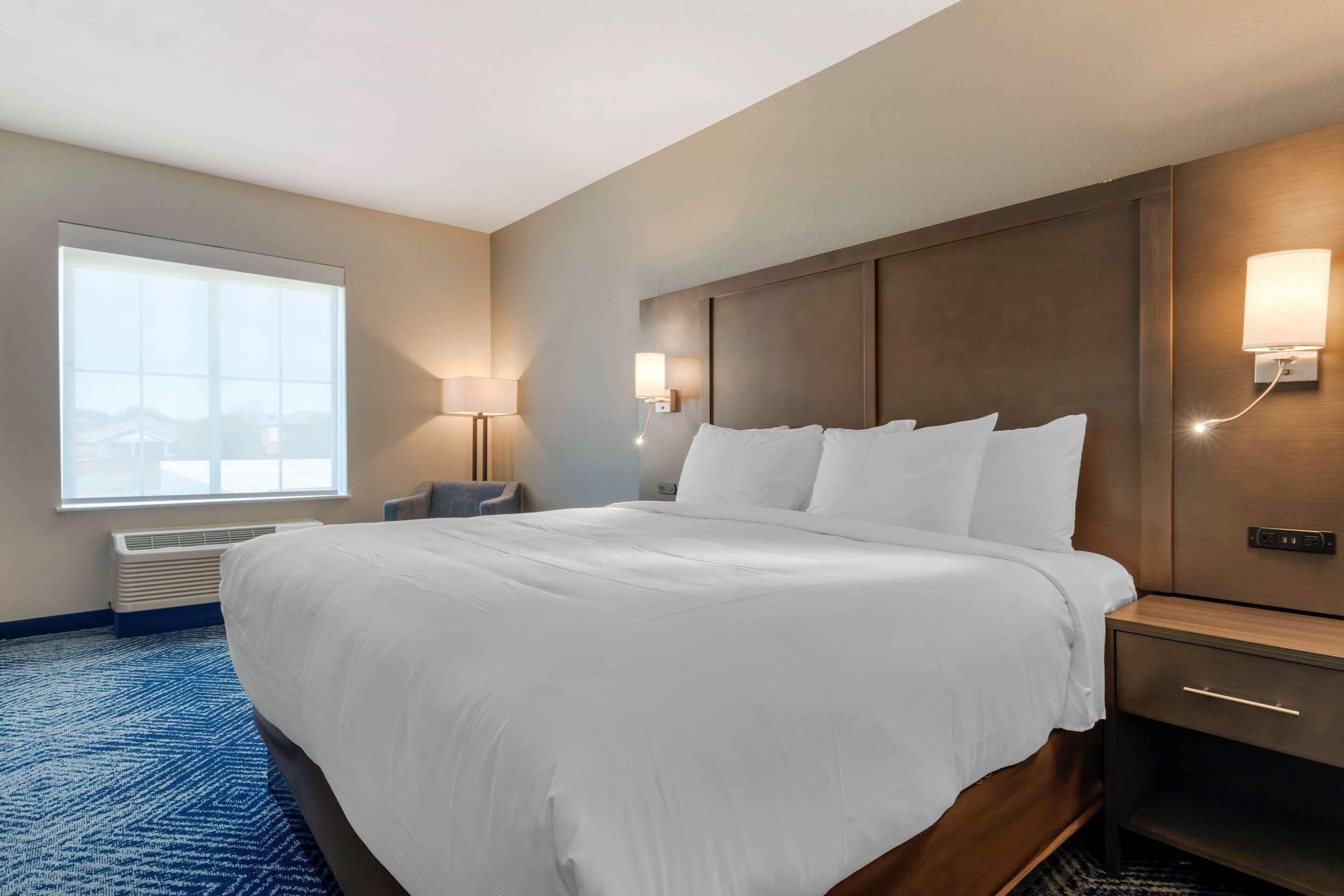 Habitación de hotel con una cama grande, una mesita de noche con lámpara, una ventana con vistas y un aplique de pared.