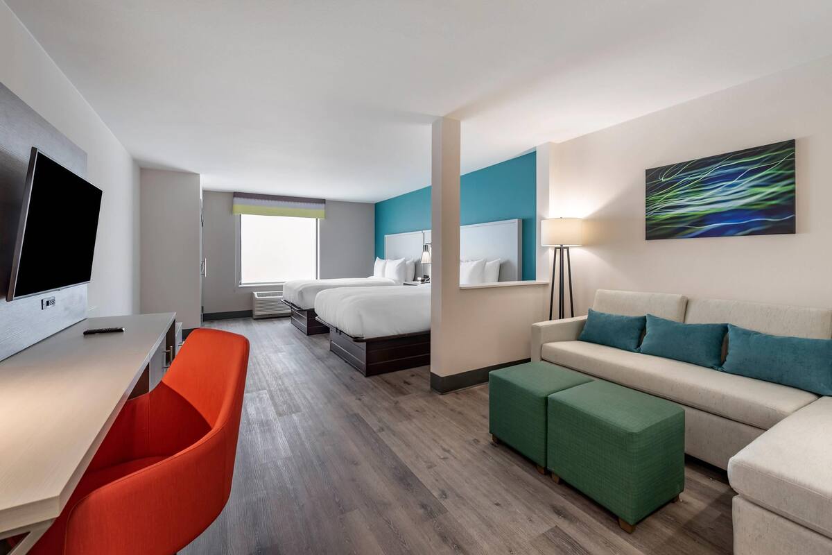 A hotel room with a bed, a desk with a TV, a sofa, and a green ottoman.