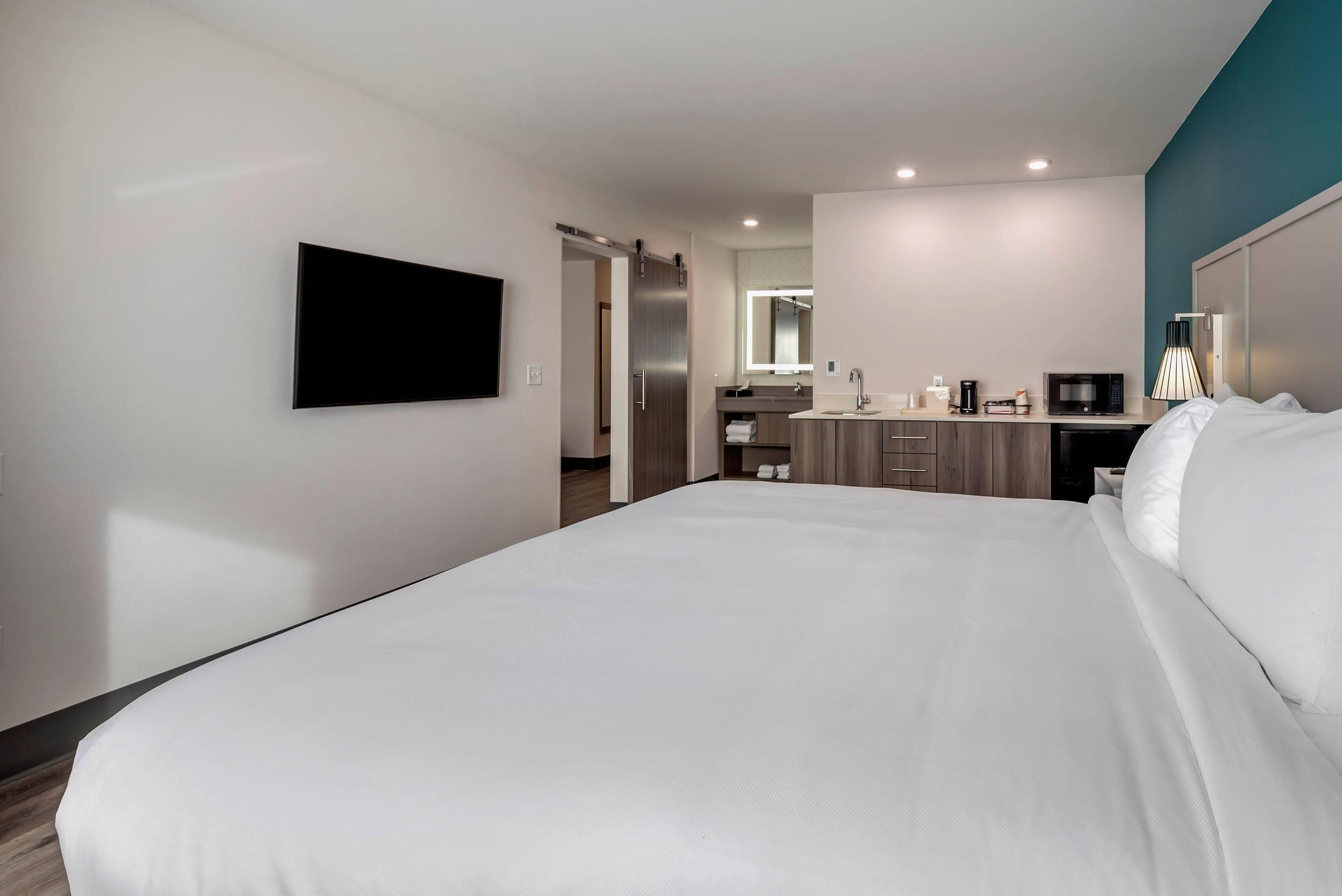 A hotel room with a large bed, a flat-screen TV mounted on the wall, a kitchenette with a microwave, and a bathroom visible through an open door.
