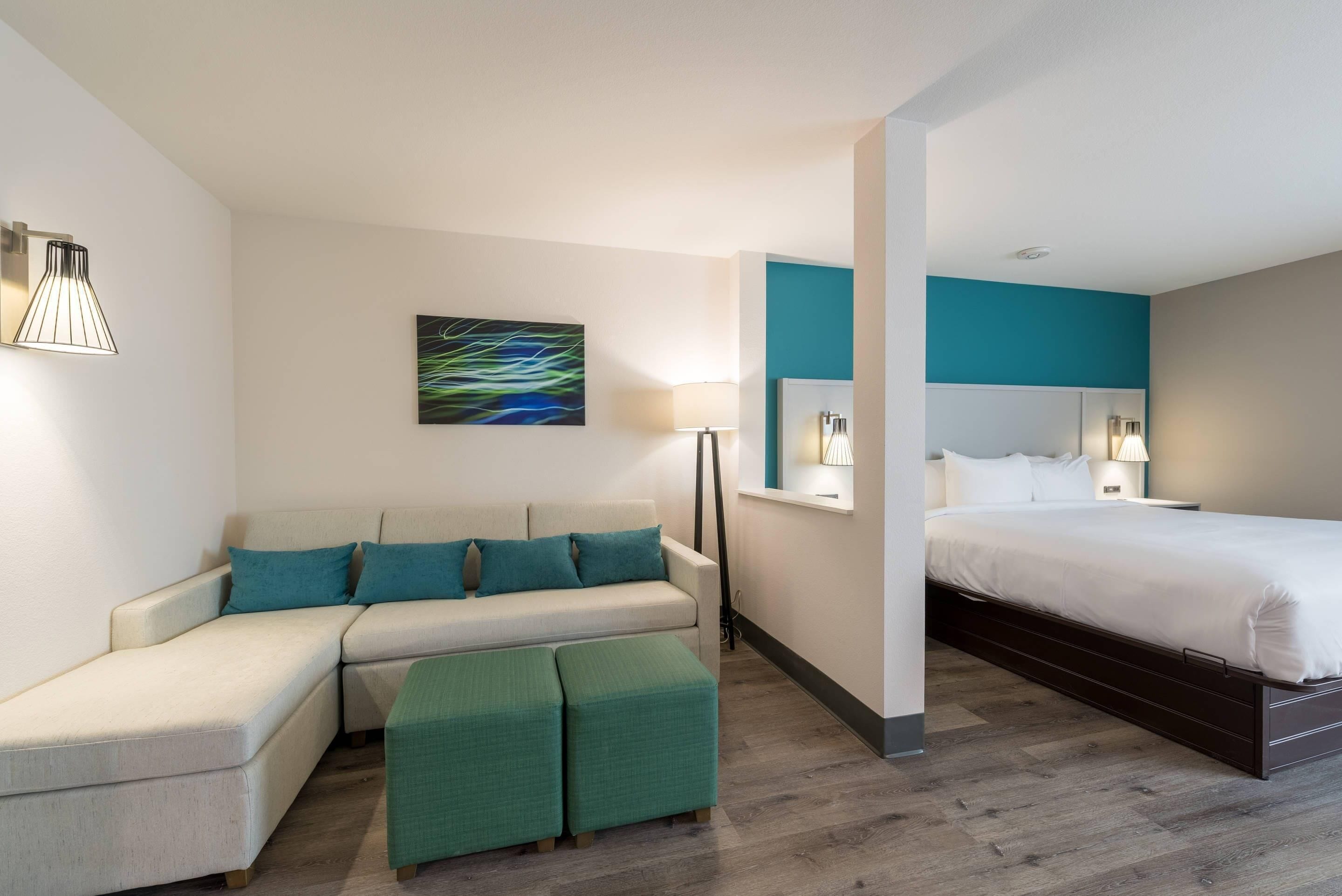 Una habitación de hotel moderna con sofá, otomanas, cama y un cuadro en la pared.