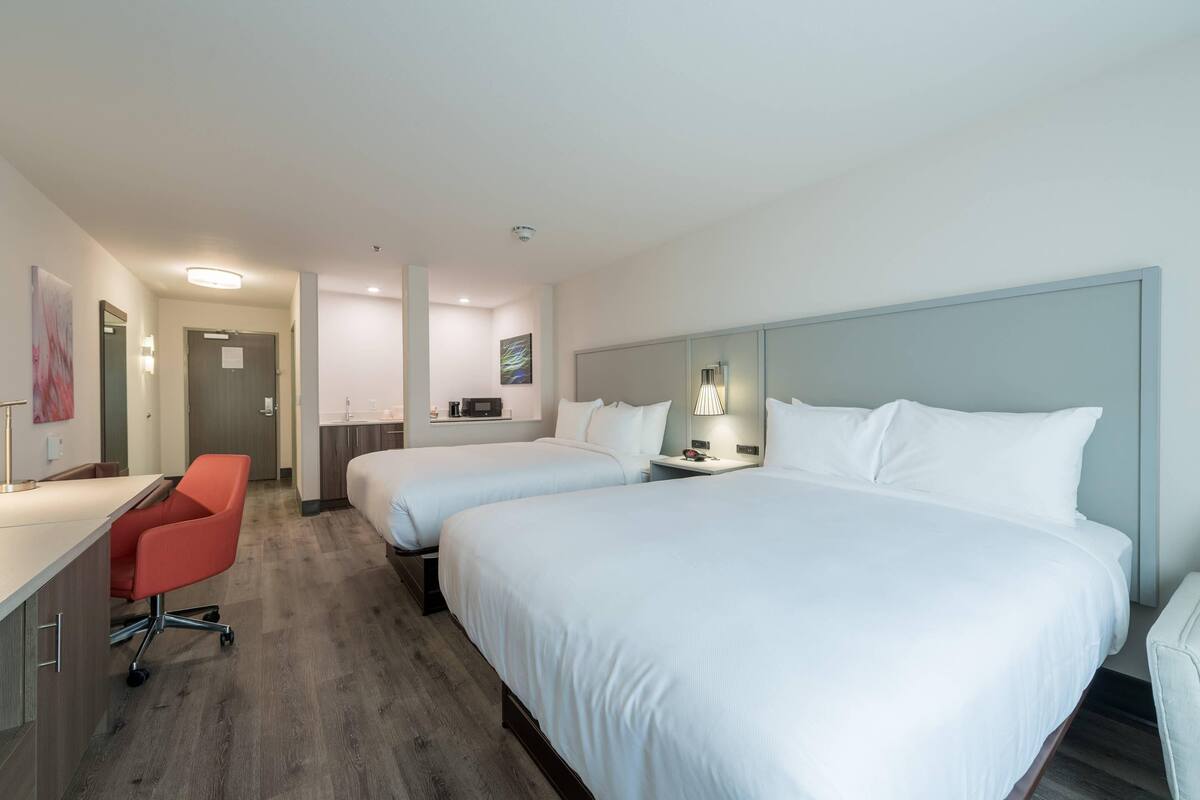 A hotel room with two single beds, a desk with a chair, a small kitchenette, and a bathroom.