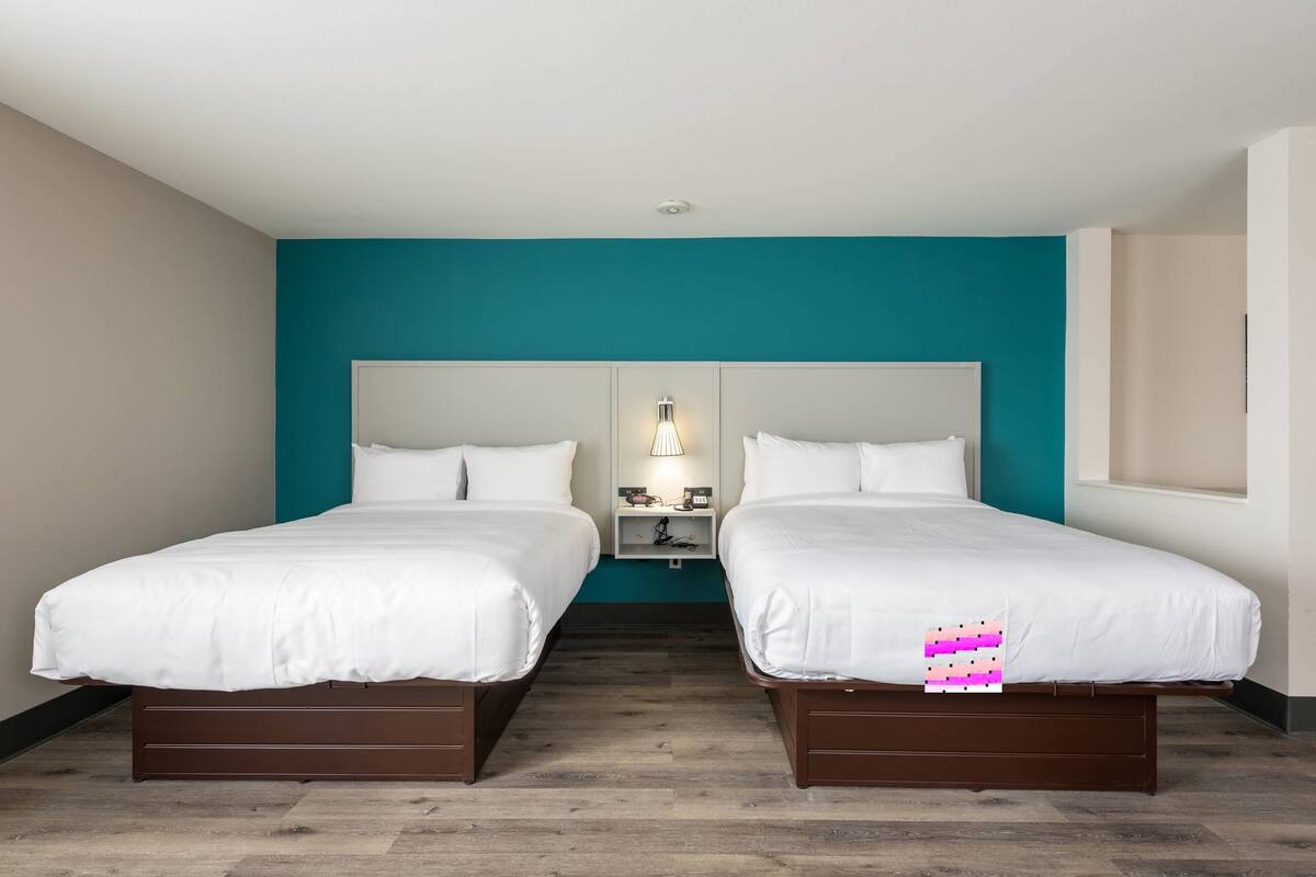 A hotel room with two beds, a teal accent wall, a white headboard, and a nightstand with a lamp.