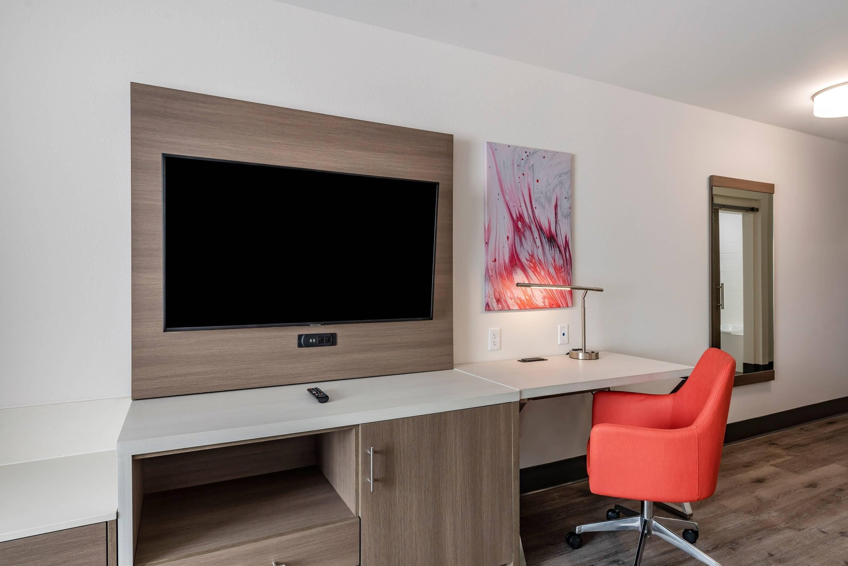 Una habitación moderna con un televisor de pantalla plana grande montado en una pared de madera, un escritorio blanco con una silla de oficina roja y un cuadro abstracto minimalista.