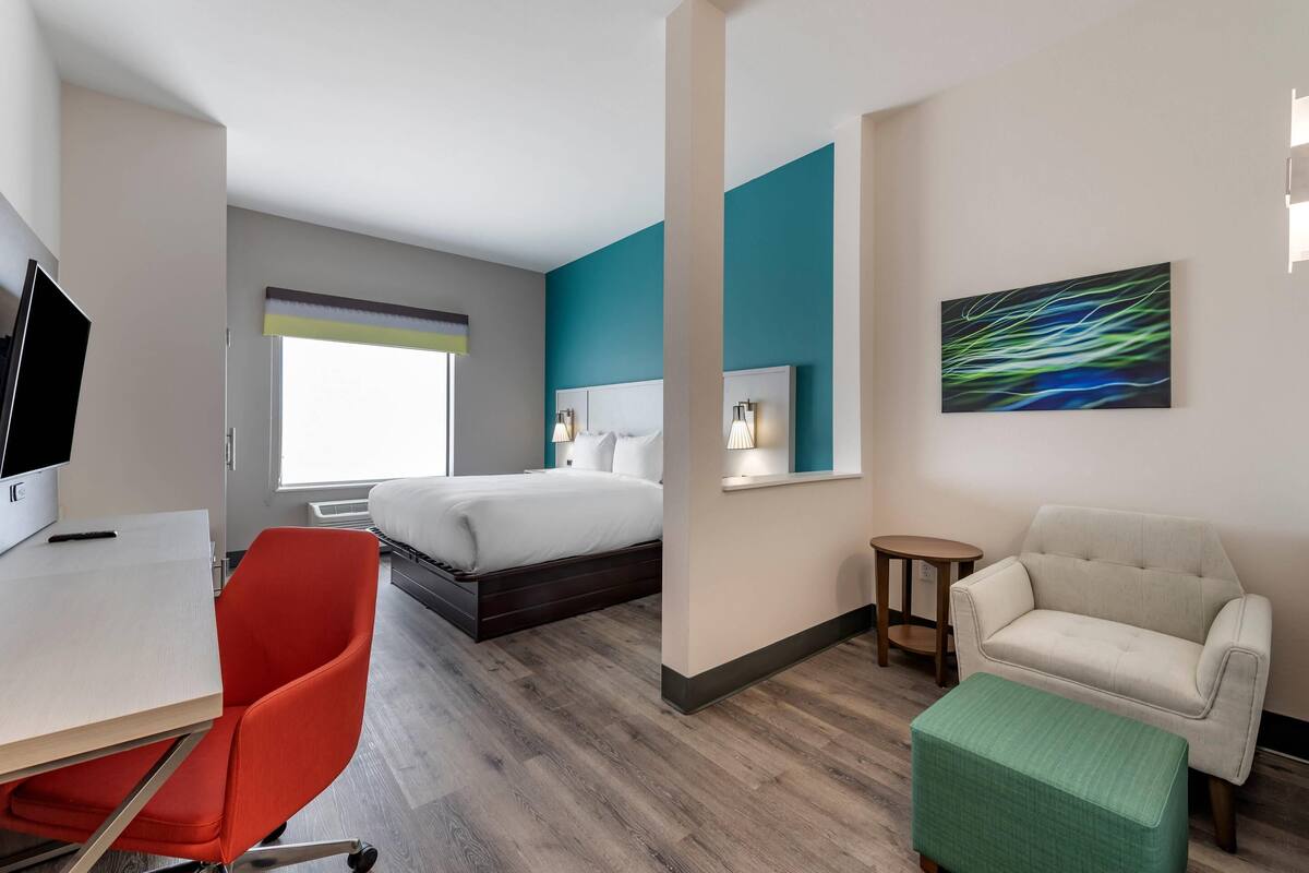 A hotel room with a bed, a desk, a red chair, a green ottoman, a white sofa, a small table, and a painting on the wall.