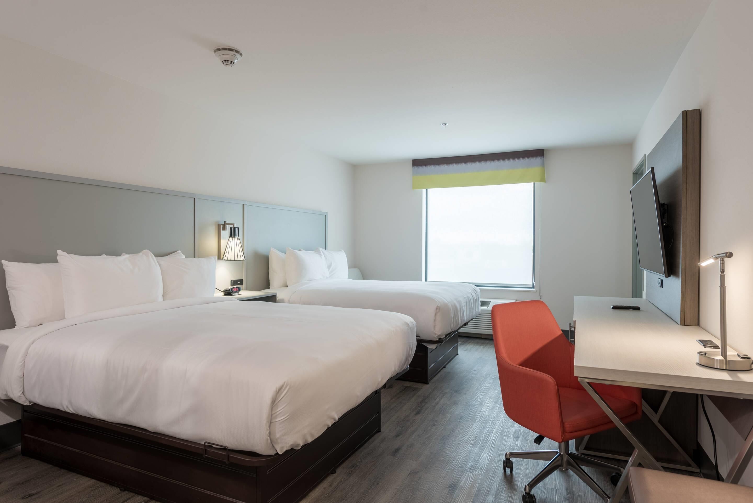 A hotel room with two beds, a desk, a chair, a TV, and a window.