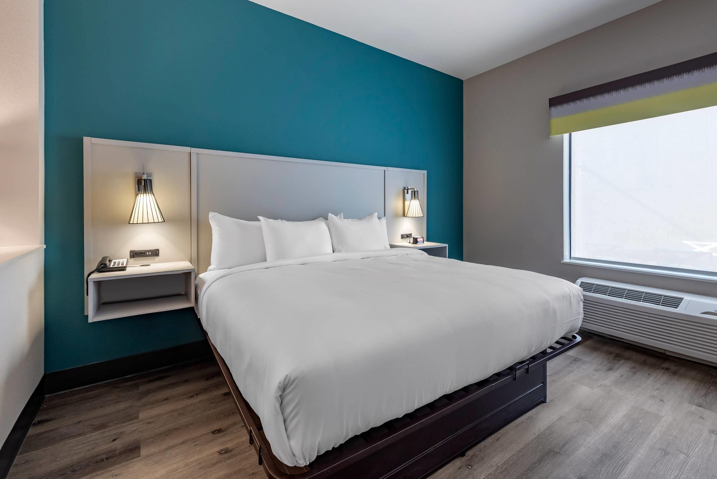 Una habitación de hotel moderna con una pared de acento color verde azulado, una cama grande con ropa de cama blanca, dos mesitas de noche con lámparas y una ventana con vistas.