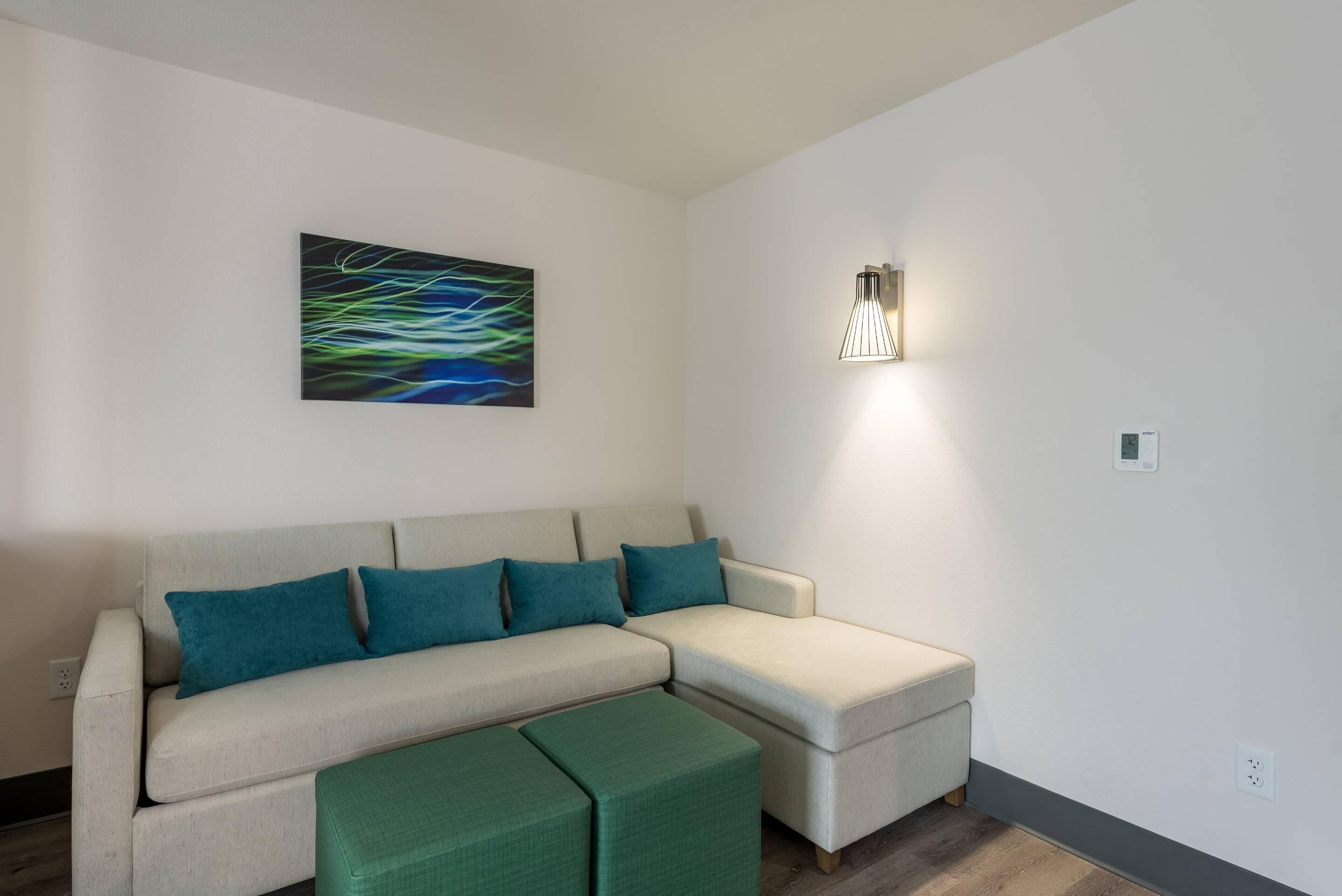 Een moderne woonkamer met een hoekbank, een groene poef, een wandlamp en een kleurrijk abstract schilderij.