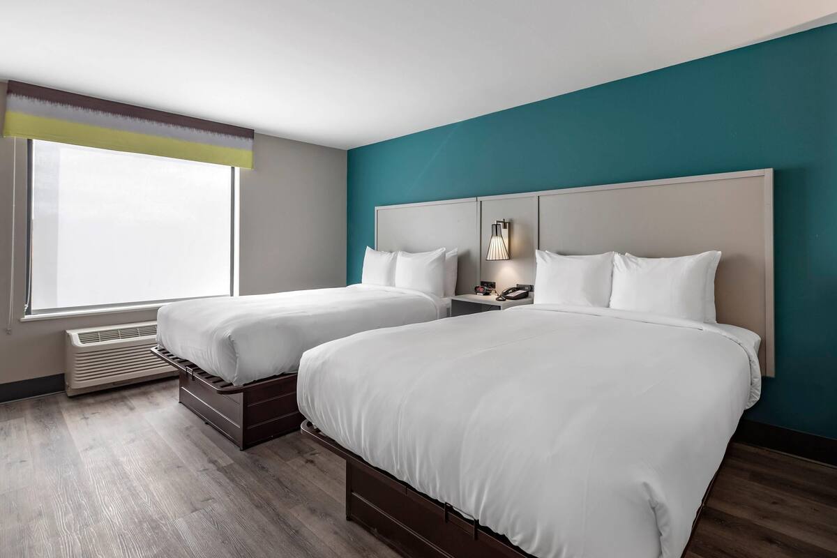 A hotel room with two beds, a window with a view, a nightstand with a lamp, and a wall-mounted phone.