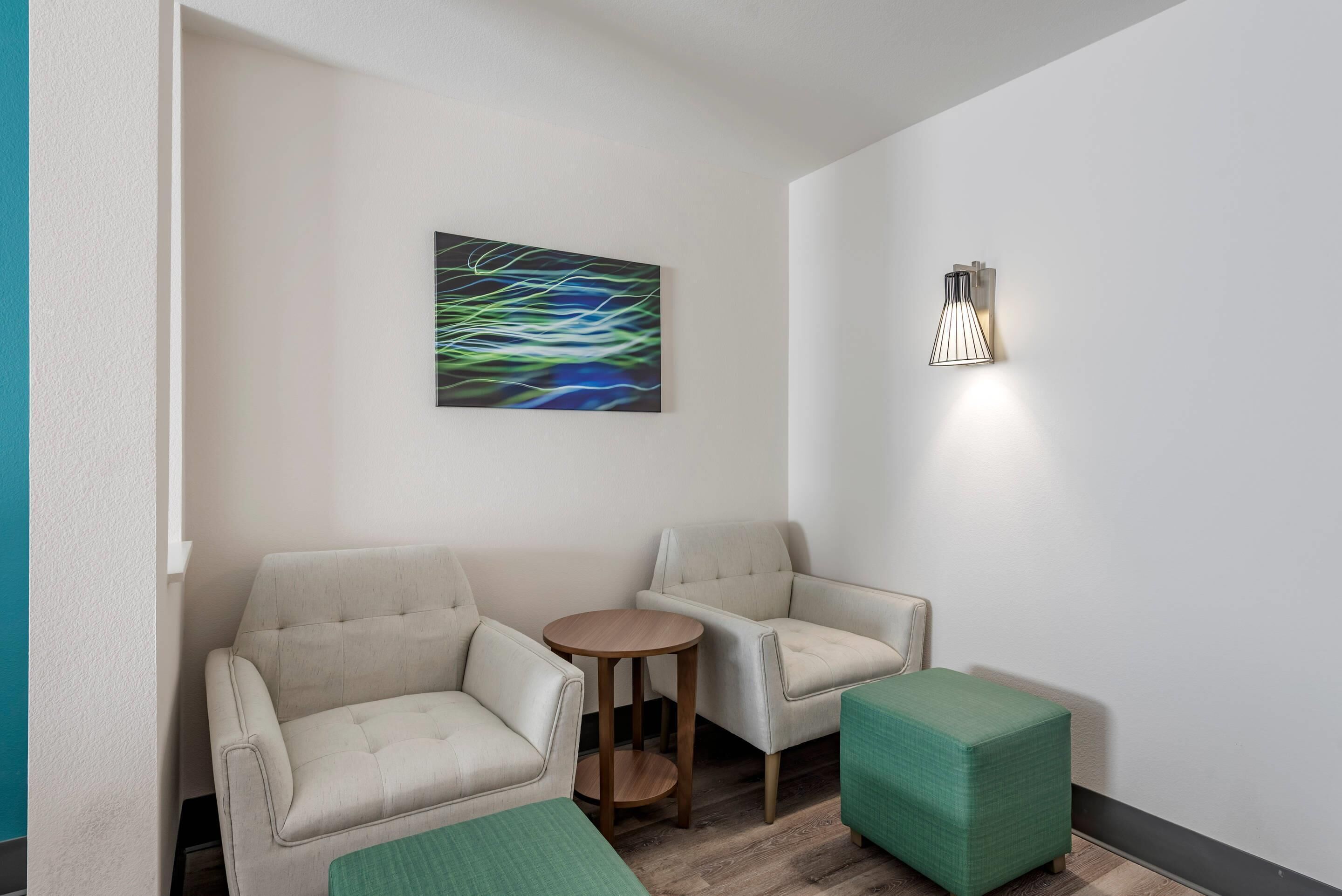 Ett modernt rum med två soffor, ett litet runt bord och en grön pall. En väggmonterad lampa och ett abstrakt konstverk syns också.
