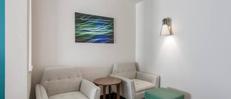 Ett modernt rum med två soffor, ett litet runt bord och en grön pall. En väggmonterad lampa och ett abstrakt konstverk syns också.