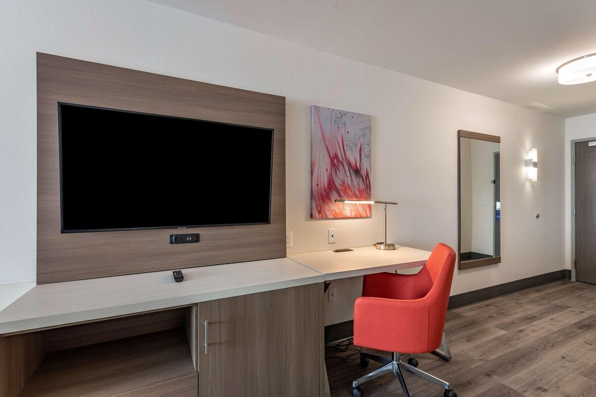 Une chambre d’hôtel moderne dotée d’un grand téléviseur à écran plat fixé au mur, d’un bureau avec une chaise et d’un grand miroir.