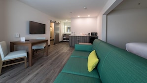 A compact hotel room with a green sofa, a wooden dining table with chairs, a flat-screen TV mounted on the wall, and a kitchenette area. - Comfort Suites Colorado Springs East -Medical Center Area (Colorado Springs)