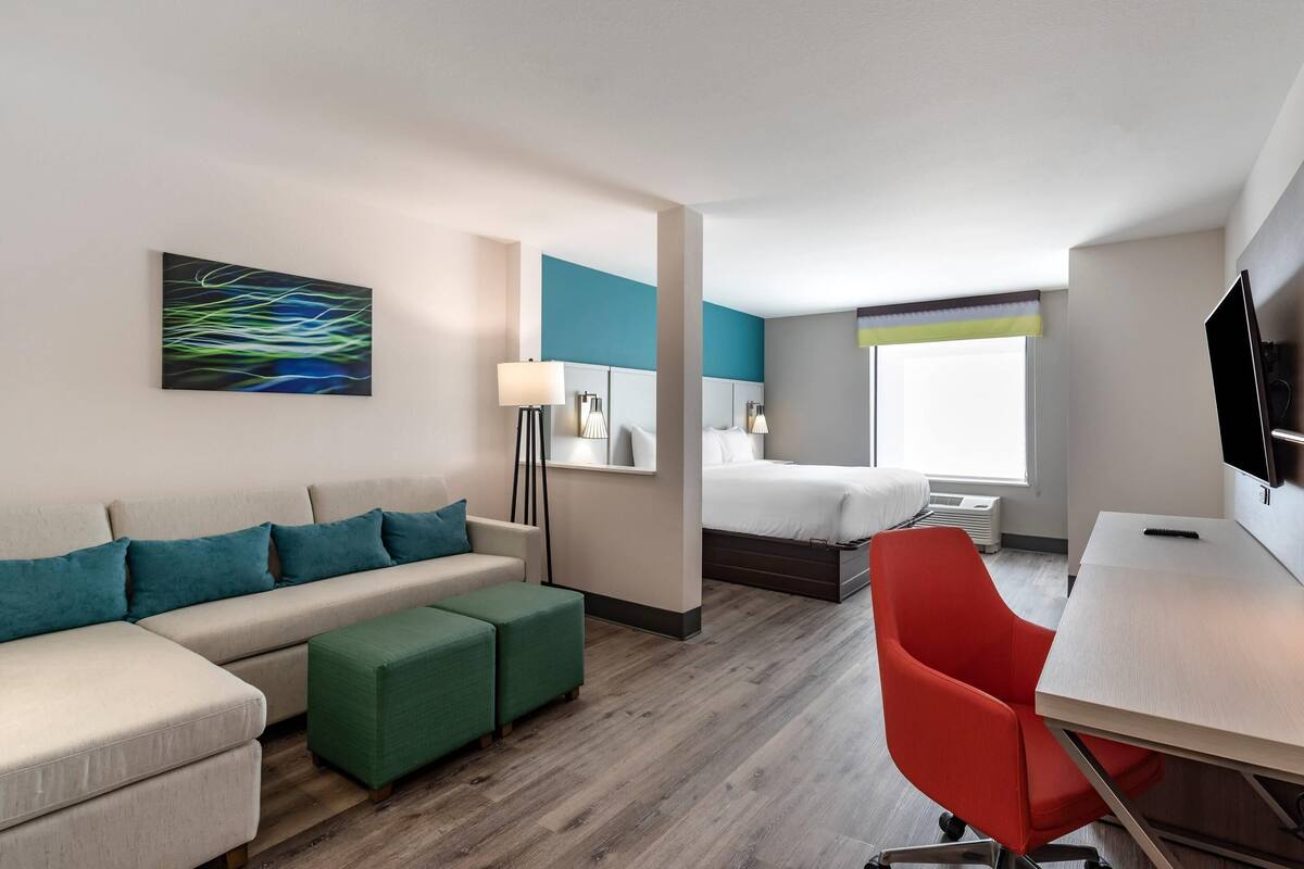 A modern hotel room with a sofa, ottoman, bed, desk, and a flat-screen TV.