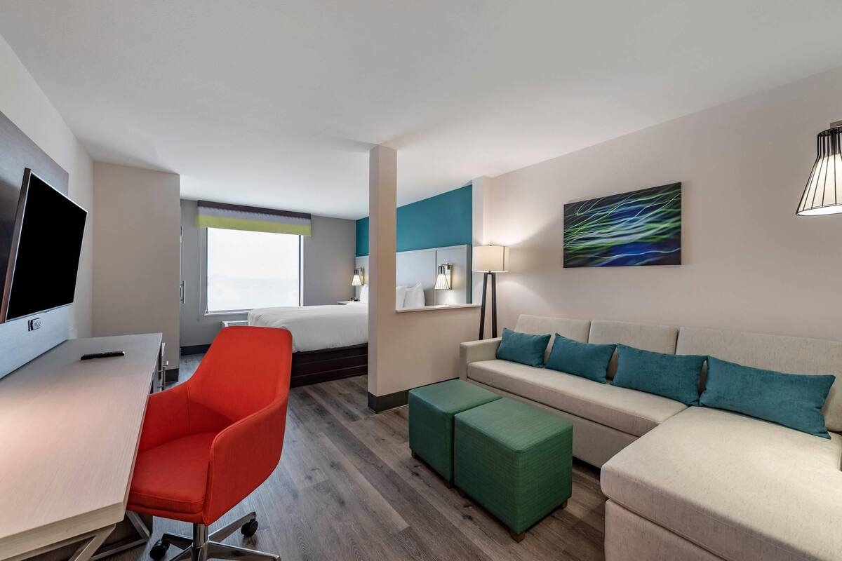 A hotel room with a bed, a desk with a computer, a red chair, a green ottoman, a beige sofa with teal cushions, a painting on the wall, and a window with a view.