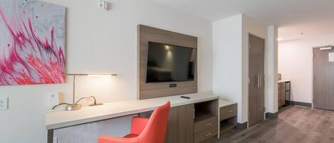 A modern hotel room with a large flat-screen TV mounted on the wall, a desk with a chair, and a wooden wardrobe.