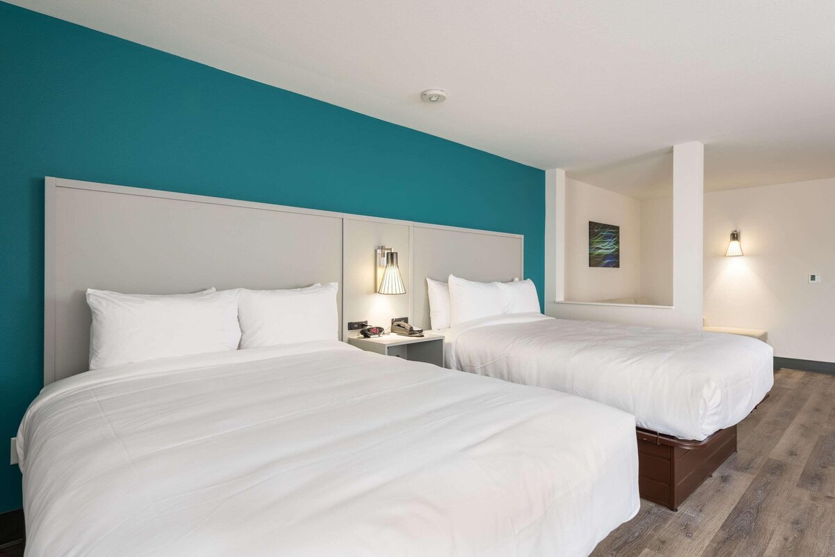 Quarto de hotel com duas camas, roupa de cama branca, paredes verde-água e luminárias modernas.