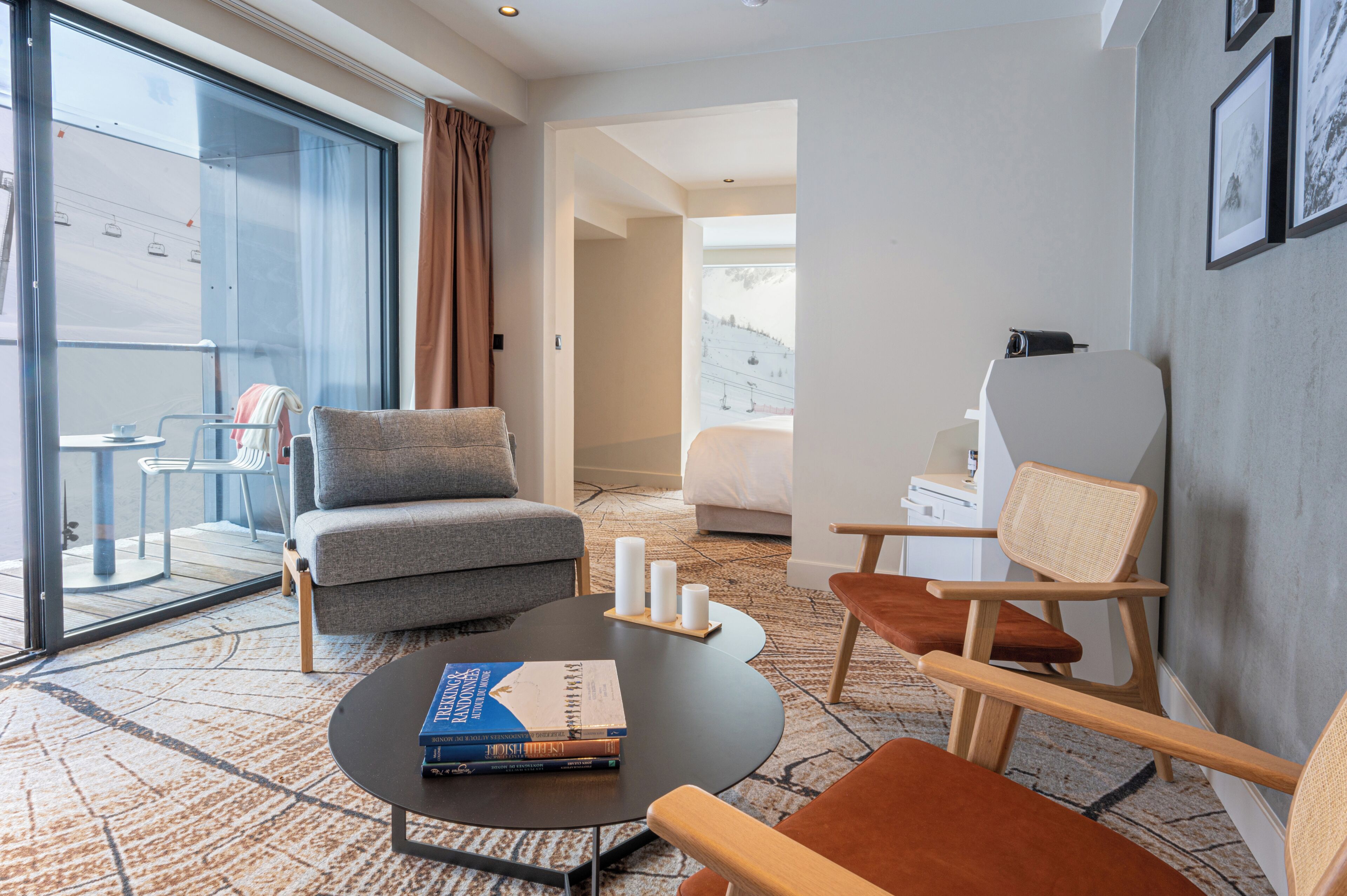 A modern hotel room with a sofa, armchairs, a coffee table with books, a bed, and a view of a ski lift.