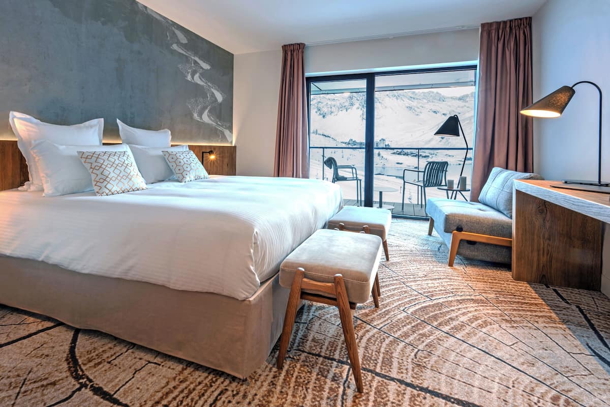 A modern hotel room with a large bed, a desk, a chair, and a view of a snowy landscape.