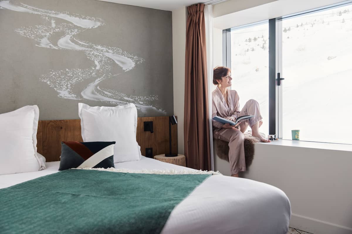 A hotel room with a bed, a window with a view, and a person sitting by the window.