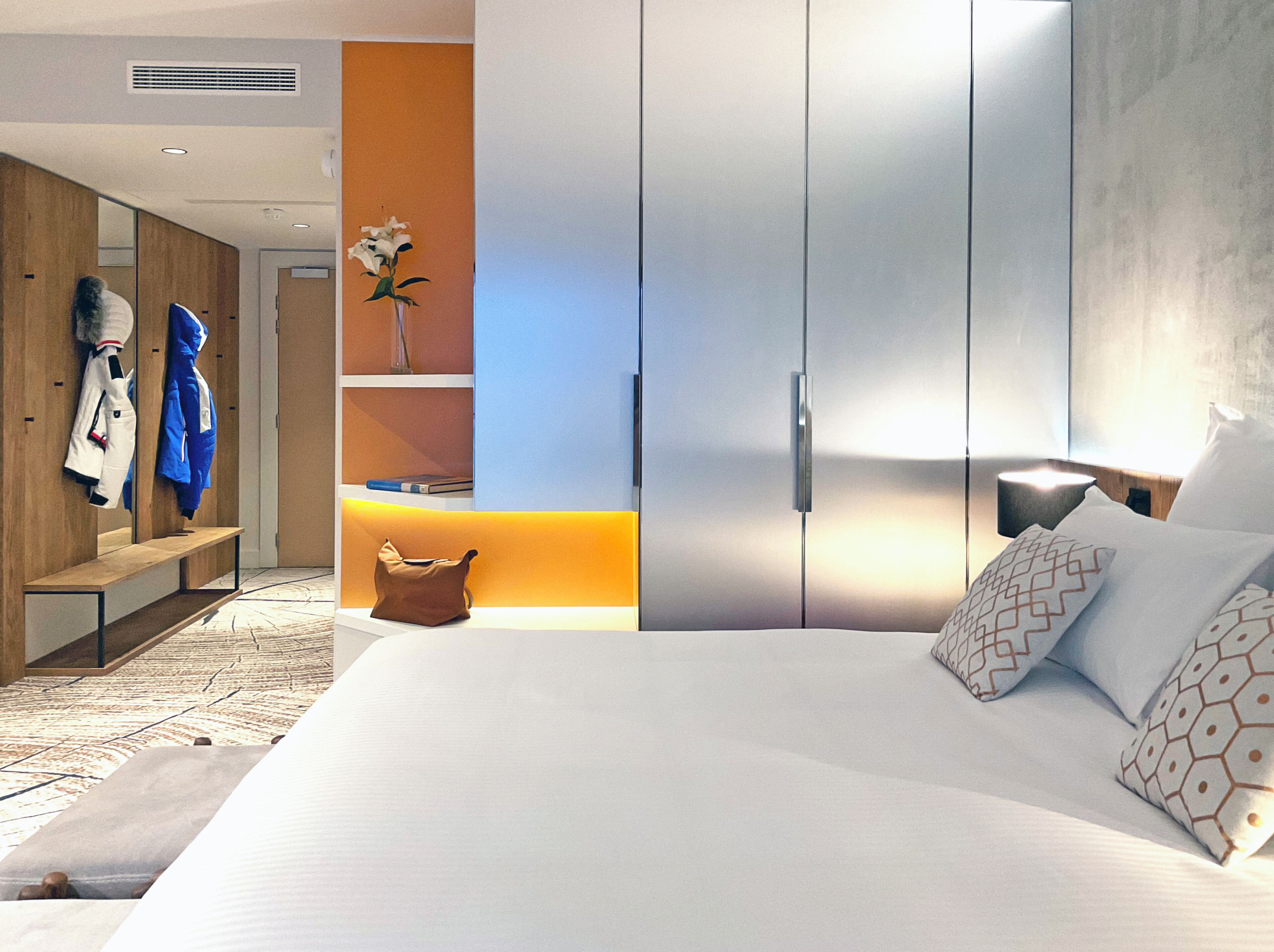 A modern hotel room with a large bed, a wardrobe, a seating area with coats, and a vase with flowers.