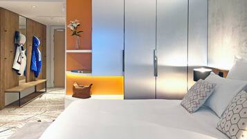 A modern hotel room with a large bed, a wardrobe, a seating area with coats, and a vase with flowers.