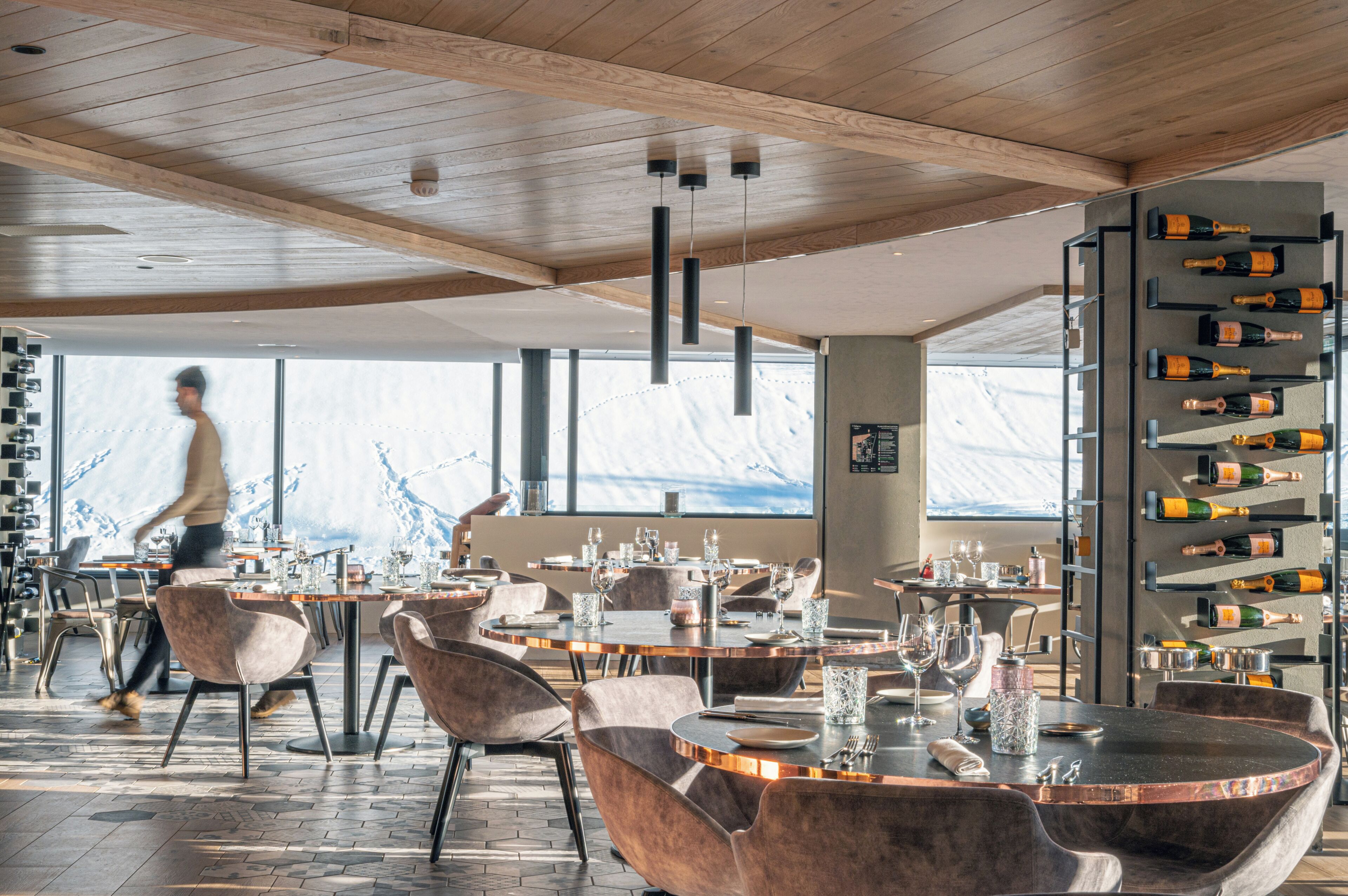 A modern restaurant interior with wooden ceiling beams, round tables, and a wine rack.