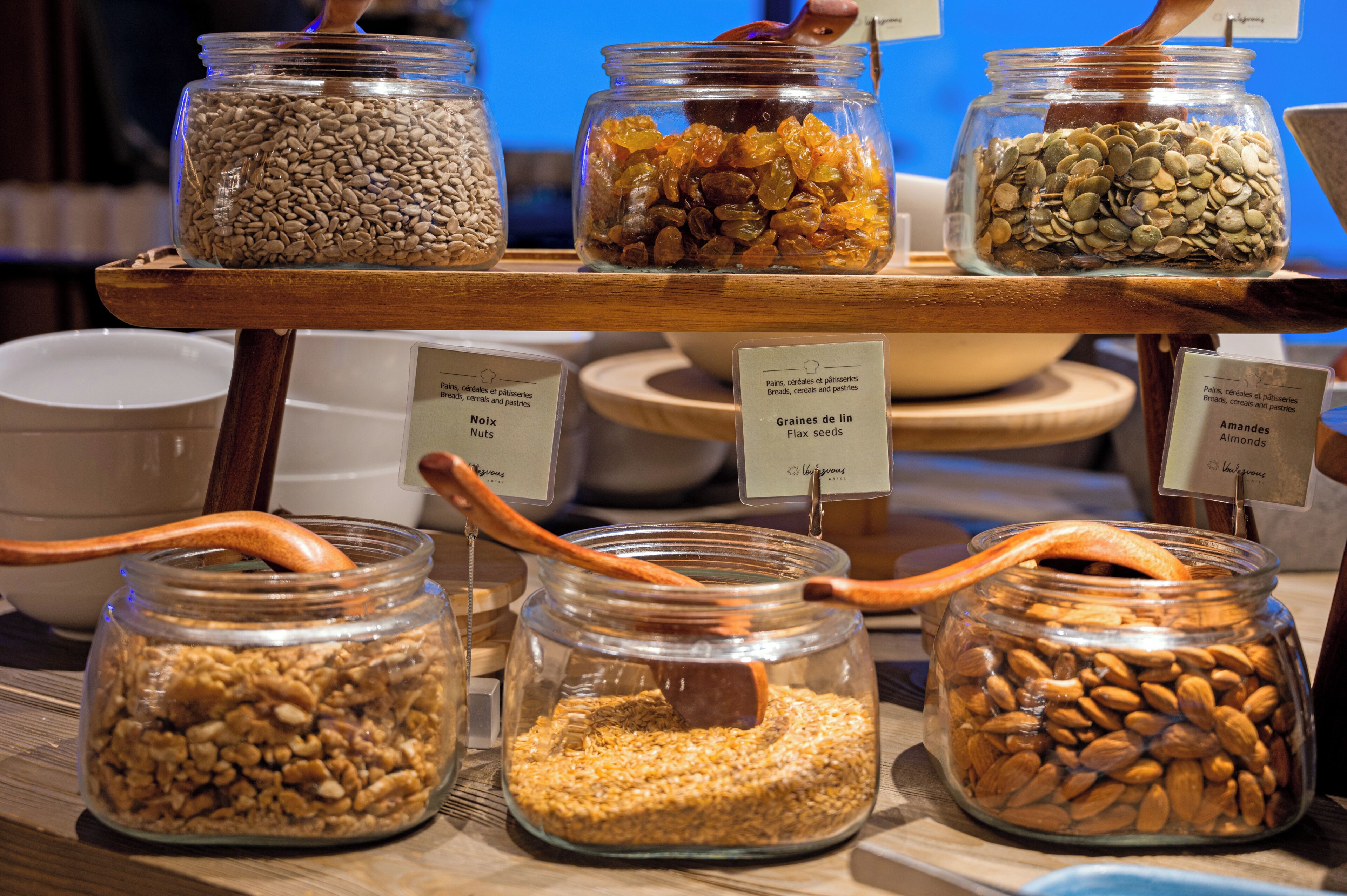 Une présentation de divers fruits à coque et graines dans des bocaux en verre, accompagnés de cuillères en bois, chaque contenant étant étiqueté pour indiquer le type de produit.