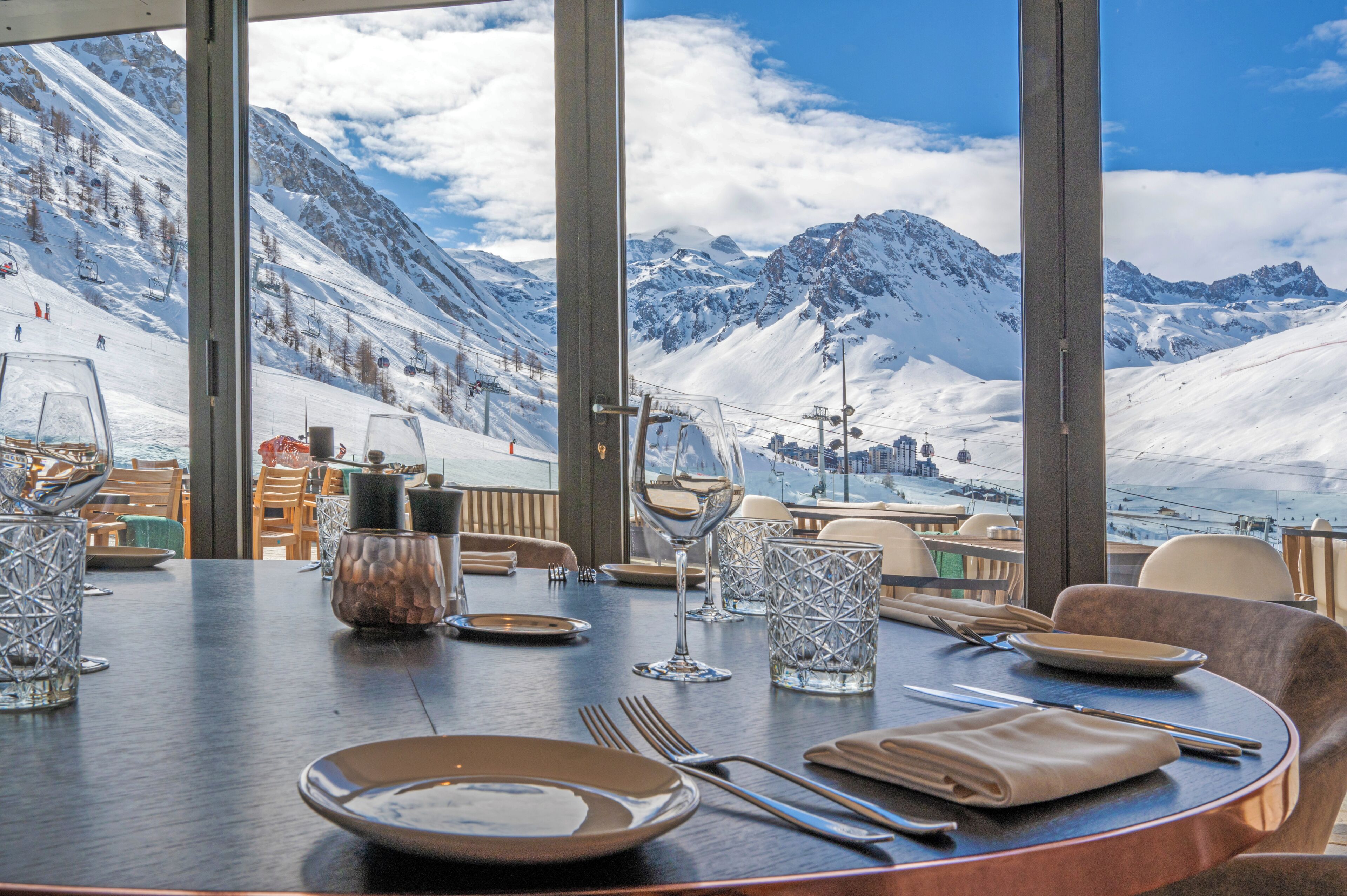 A dining table set with plates, glasses, and cutlery, overlooking a snowy mountain landscape with a ski lift in the distance.