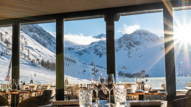 Un restaurante con vistas a la montaña, mesas dispuestas con copas de vino y un telesilla al fondo.