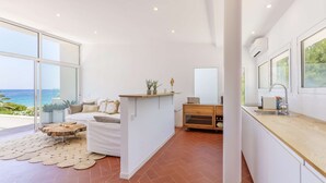 Una sala de estar moderna con un ventanal que da al mar, un sofá blanco, una mesa de centro de madera y una cocina con un fregadero y encimera.