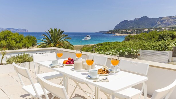 Una terraza con mesas y sillas blancas, una mesa dispuesta con zumo de naranja, frutas y bollería, con vistas a una zona costera con un barco y montañas.